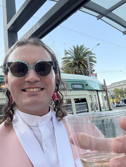 An enby wearing a pink suit, blue glasses, and Muni T Third earrings holding a plastic cup of white wine in front of a streetcar with green and cream livery