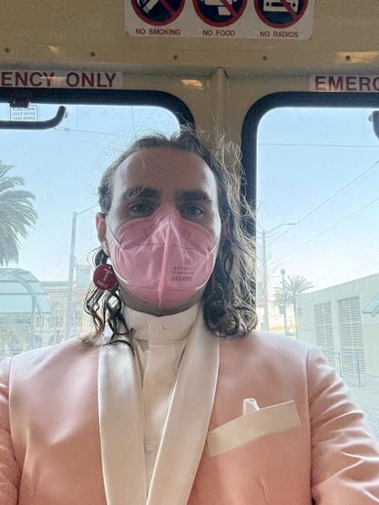 An enby wearing a pink suit and pink mask sitting in the back of a streetcar