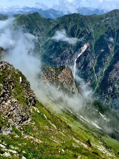 Photo of mountains in the background and a a rock and steep slope in the foreground. There are clouds in the steep slope.