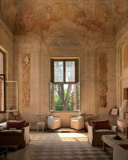 High ceilings covered in italian murals in golden and orange colours. Windows with rays of sunshine coming through. Light furniture is spread around, leaving the spotlight to the mural.