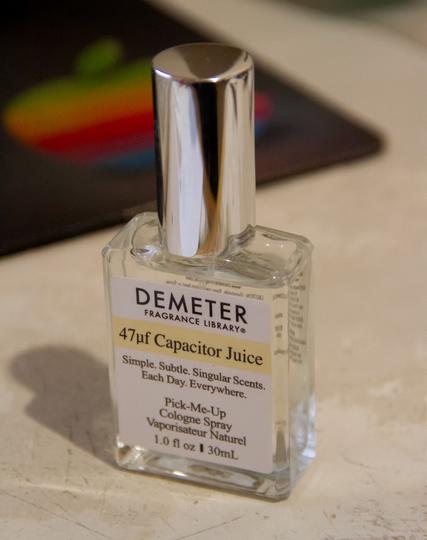 A photo of a bottle of a Demeter fragrance on a desk, with an Apple mouse pad behind. This one is the scent '47uf Capacitor Juice".