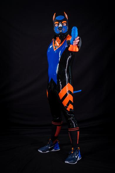 Human pup wearing Black, Royal Blue, and orange full rubber catsuit with a blue tail plug visible behind him. He's looking happy and holding an orange and blue dildo out towards the viewer