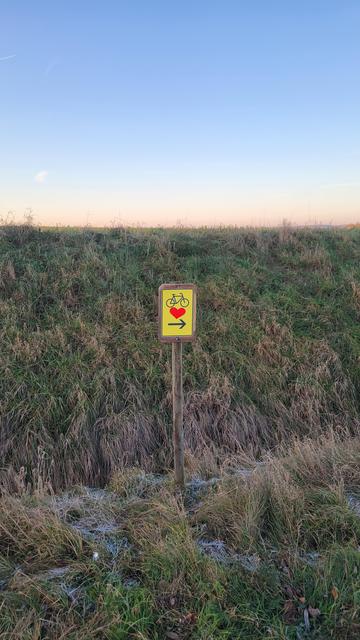 Ein gelbes [inoffizielles] Schild mit einem Fahrrad, Herz und Pfeil am Wegrand zeigt die Richtung an.