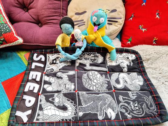 Three sock toys are sitting together under a small black quilt printed with D and D monsters and the word "sleepy" on the side. There is a large skeleton, a small skeleton, and a zombie. The sock toys are wearing homemade winter clothes.