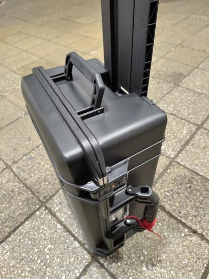 A black flight case sitting on a station floor, handle extended, a small red ribbon tied around one handle.