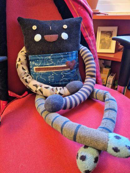 A handmade stuffed toy of a robot made from recycled fabrics. He has long limbs made of socks with little round hands and feet. His body is made of soft corduroy. He has button eyes and a happy smiling face. On a chest is a heart meter which is full.