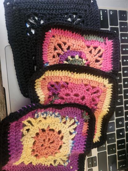 Photo showing 4 crochet squares using the Emma square pattern. One is all black, 3 are multicoloured (lots of yellow, orange, pink, purple, and a little bit of blue, white, and green), with a black border. Squares are overlapping and are set on an aluminium laptop keyboard.