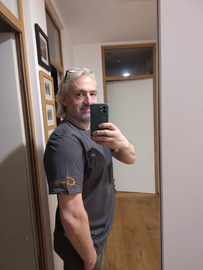 Selfie leines Mannes im Spiegel mit einem mit Firmenlogo bedrucktem T-Shirt im Hintergrund ein Flur mit Eichenparkett und weißen Zinmertüren mit Glasoberlichtern, Bilder an einer Wand.
