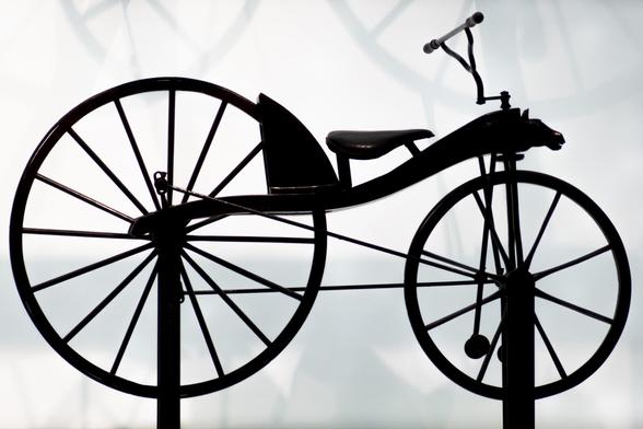 Photo of an old-fashioned bicycle, with a big back wheel and small front wheel. Photo is in silouette against light gray background.