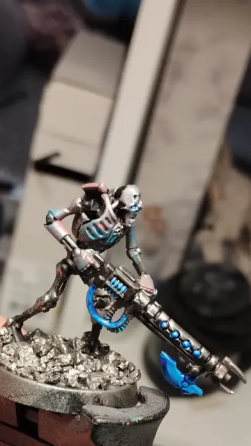 Necron warrior with a black gauss flayer looking to the right.
It has a metallic armor which turns from blue to pink.
The energy highlights on the weapon are a bright blue.