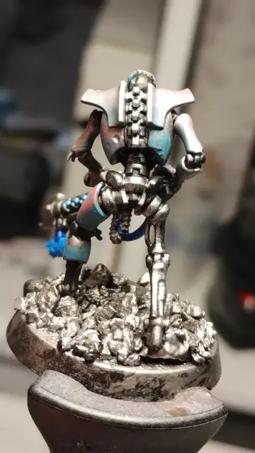 Necron warrior with a black gauss flayer from behind.
It has a metallic armor which turns from blue to pink.
The energy highlights on the weapon are a bright blue.