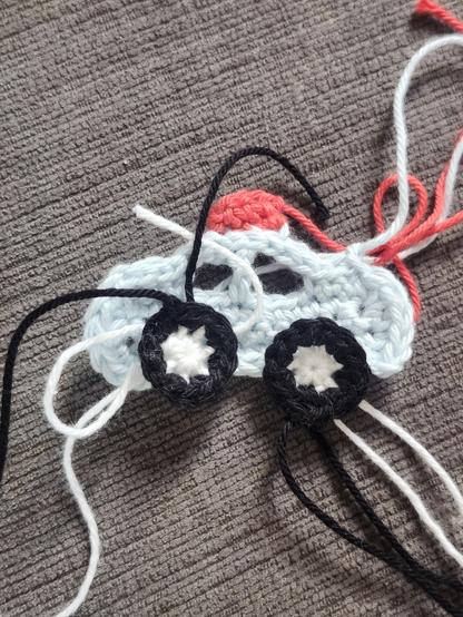 2 dimentional crocheted piece of light blue (almost white) police car with black tires and a red light on top. It’s viewed from the side, and it’s laying on top of grey fabric. About a inch and a half in width. It’s an applique made to be sewn on a crocheted crown.