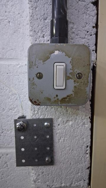 An ordinary light switch, of the grey box and conduit sort common in garages and industrial sites. Beneath it is another switch that is clearly an old panel mount switch crudely attached to the wall with a scrap metal bracket. A run of doorbell wire runs upwards through the air and out of frame.