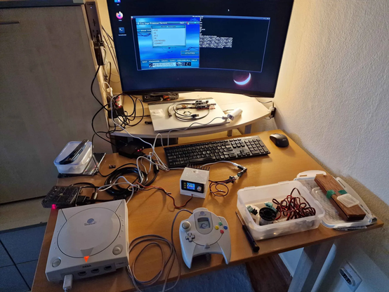 A desk full of wiring, Dreamcast, power supply, soldering iron, and similar mess. There is a monitor that shows the dreamcast's video output and a terminal with the receiving modem's raw output.