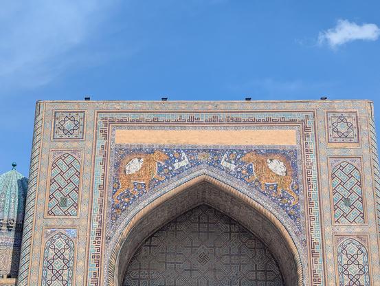 Photo du haut de la porte de la médersa (école coranique) de Cher-Dor. Au milieu, une grande mosaïque représentant sur fond bleu et fleuri des animaux fantastiques : des sortes de cerfs/biches et surtout des lions-tigres portant deux têtes entourées d'un soleil. Sur les côtes et au-dessus de la mosaïque centrale : des mosaïques représentant des motifs géométriques avec plusieurs nuances de bleu.