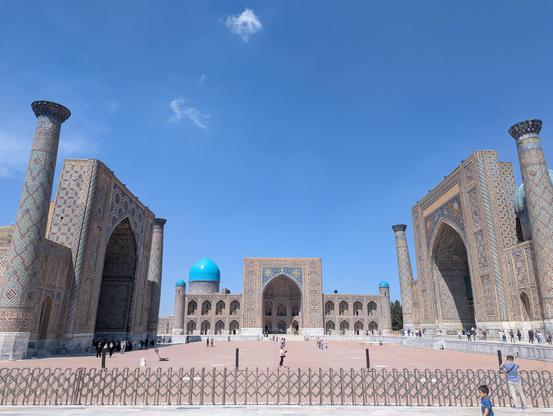 Une grande place carrée avec 3 grands bâtiments, qui sont des anciennes médersas (écoles coraniques) décorées de mosaïques dans des nuances de bleu, principalement des motifs floraux et géométriques.