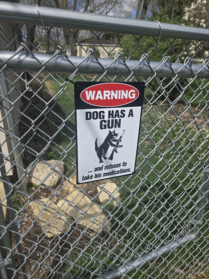 Warning sign on a chain link fence that depicts a dog with a rifle. It reads "Warning: Dog has a gun and refuses to take his medication."