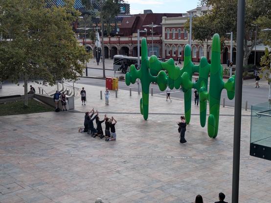 A photo of the "Grow Your Own" sculpture in Forrest Place, affectionately known as "The Cactus". In front of it, five men are in a row kneeling with their hands in the air worshipping the cactus.