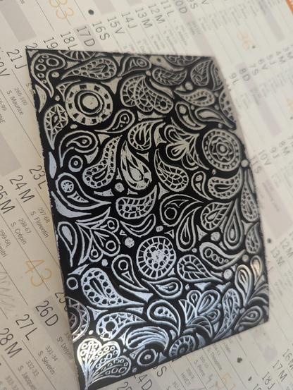 Une carte a gratter de format carte postale (10x15cm environ), a l'origine noire, est recouverte de motifs abstraits intriqués rappelant des feuilles et des fleurs. 
Le dessous de la carte à gratter est argenté.