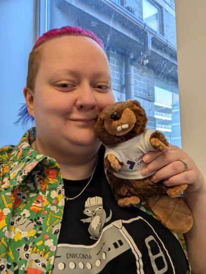 Cat, with pink, purple and blue hair, holding a Beaver. She is wearing a green shirt with trash pandas and a t-shirt with gophers on. 

Look for the colour clash.