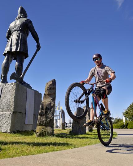 a wide shot in portrait of a park on a sunny day. The sky is blue and cloudless with green grass and a path the leads towards the camera. On the left side a statue holding an axe is seen from behind. on the right side a bicycle is doing a wheelie toward the right.
