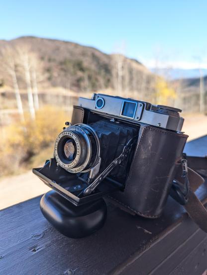 A folder camera with the bellows open and a half leather sleeve protecting the sides and providing attachment for the leather straps is propped up on a balcony railing with a sunny hillside out of focus in the background.