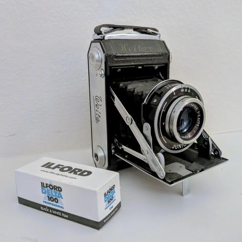 Color photo of an old fashioned folding bellows camera and a roll of 120 film.