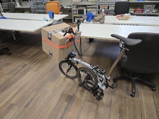A Brompton folding bike parked inside with a big cardboard box strapped to the front carrier