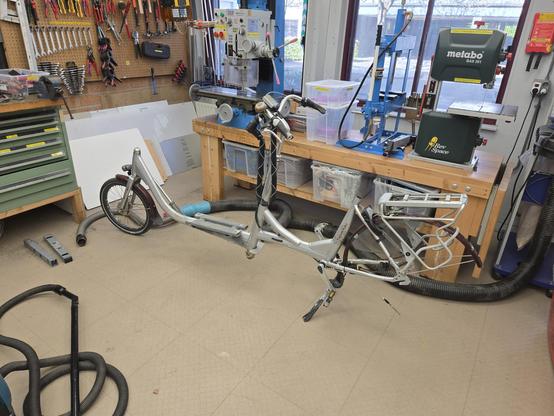 A cargo bike sitting in a workshop. The tub in the front is missing, so it looks more like a stretched out normal bike. Also, it has no rear wheel.