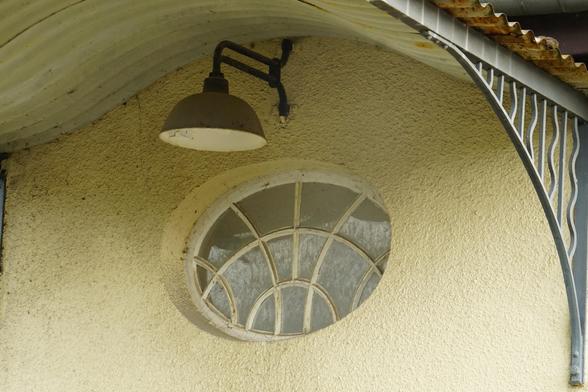 ein kleines Ovales Fenster mit vielen gebogenen Sprossen in einer gelb/weisen Wand. Darüber eine alte Lampe und gerade noch zu erkennen ein gebogenes Blechdach.