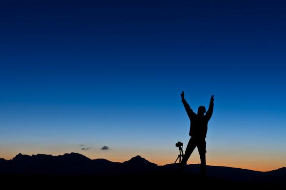 Photo d'un personnage en ombre chinoise sur fond de coucher de soleil en montagne.
Le personnage lève les bras en signe de victoire, devant lui on voit un appareil photo reflex sur trépied. La ligne d'horizon, tout en bas de l'image, est montagneuse. Le ciel est orangé au raz de l'horizon, et passe au bleu nuit assez rapidement à mesure que l'on regarde vers le haut de la photo.