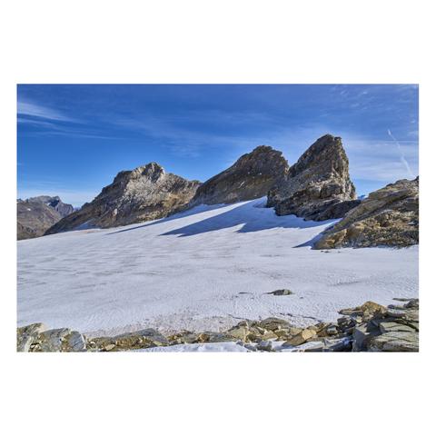 Photo d'un paysage de haute montagne. 
On voit trois pics rocheux devant un petit plateau glaciaire bien blanc. L'ambiance est minérale, le ciel est bleu malgré de légers voiles nuageux.
-
Photo of a high-mountain landscape. 
Three rocky peaks are visible against a small, snow-white glacial plateau. The scene has a rugged, rocky feel, and the sky is blue despite a light veil of clouds.