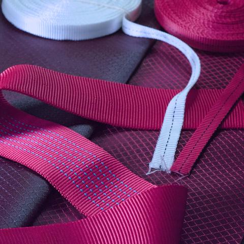 a few layers of fabric arranged to compare how the colors look next to each other. The base layer is red and black diamond ripstop with a wide band of red tubular webbing laid over it and two thin straps of red and white webbing on top for comparison