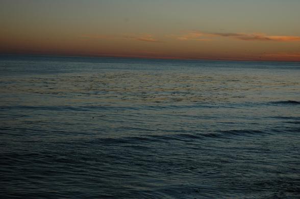 Eine Landschaftsaufnahme des Meeres während der Abenddämmerung. Der Himmel zeigt einen Farbverlauf von einem dunklen Blau am oberen Rand zu einem warmen Orange am Horizont. Einige dünne, horizontale Wolkenstreifen sind im Himmel sichtbar. Das dunkle Wasser des Ozeans weist sanfte, wellenförmig wellenförmige Bewegungen auf. Auf der Wasseroberfläche sind leichte Lichtreflexionen erkennbar.