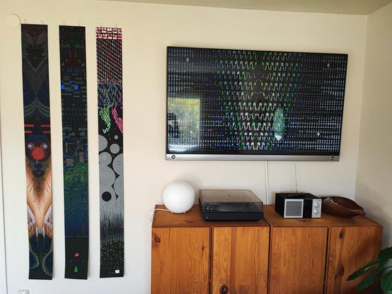 A photo of three prints hanging to the left on a wall. On the right is a TV-screen that shows abstract ASCII patterns.