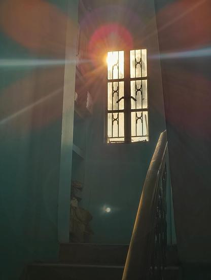 A low-angle view looking up a dark indoor staircase toward a small, multi-paned window. Intense, warm sunlight is bursting through the glass, casting dramatic, glowing light rays and large, colorful lens flares across the dim walls. A smooth wooden handrail curves up the right side of the frame, disappearing into the shadows.