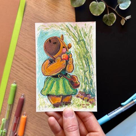 Picture of my hand holding up an original gel pen painting of a little anthropomorphic capybara wearing a green dress with a brown belt decorated with a golden heart-shaped buckle. She is wearing a little brown satchel and is standing on a grassy patch, a bunch of bamboo is planted next to her.