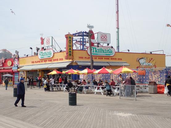 Coney Island à New York.
Devant l'enseigne au Nathans au bord de la plage
On y voit le bâtiment, avec des tables devant.
Chaques tables ont des parasols rouge et jaune.
On y voit aussi un passant marcher devant la camera sur les planches de la promenade