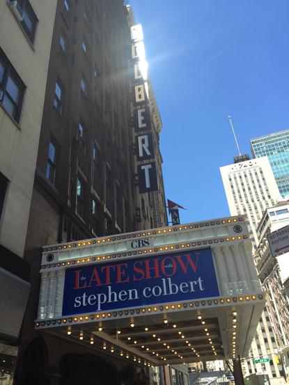 The marquee of the Ed Sullivan Theater with text “The Late Show with Stephen Colbert” and giant letters “COLBERT” on the vertical part of the marquee. The theater is photographed from the ground looking up, against bright blue summer sky and nearby buildings In Manhattan.