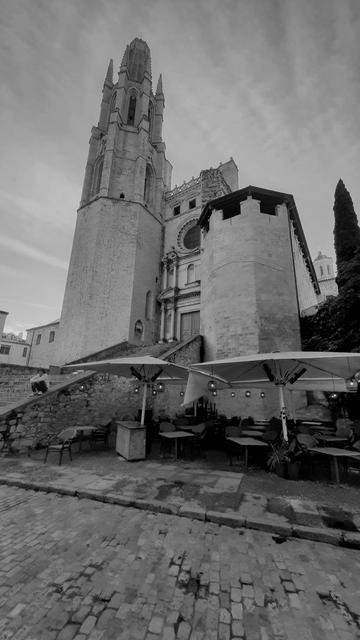 Picture showing a church with rather prominent perspective in blackandwhite. In rhe foreground stars with a young man sitting p  the rail, and some sunshades and Tabletten of a restaurant nearby