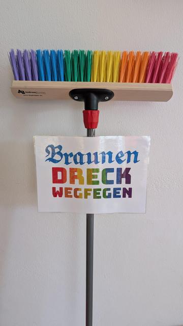 Besen in Regenbogen-Farben mit Schild "Braunen Dreck wegfegen"