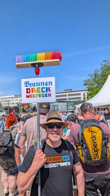 Mann mit Regenbogenbesen und Schild mit Aufschrift "Braunen Dreck wegfegen"