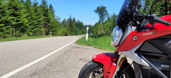 A long straight road in a Forrest on a sunny day. In the background two windmills can be seen. The right side of the picture is a red electric motorcycle facing the road.