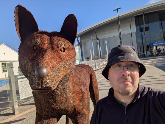 A selfie of me next to a large statue of a dog. I am wearing a black bucket hat and purple fleece sweater. The dog statue's head is about twice as large as mine, made of rusted iron. In the background is the WA maritime museum.