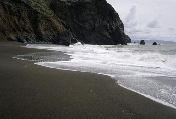 Eine Küstenlandschaft mit einem dunklen Sandstrand, steilen Klippen und Wellen. Im Vordergrund befindet sich dunkler, fast schwarzer Sand, der von weißer, schäumender Brandung überspült wird. Auf der linken Seite ragen große, dunkle Felswände auf, die teilweise mit grüner Vegetation bedeckt sind. Im Hintergrund sind weitere kleine Felsformationen im Meer unter einem bewölkten, grauen Himmel zu sehen. Die Szene zeigt den Übergang zwischen dem Strand, dem Meer und den Klippen.
