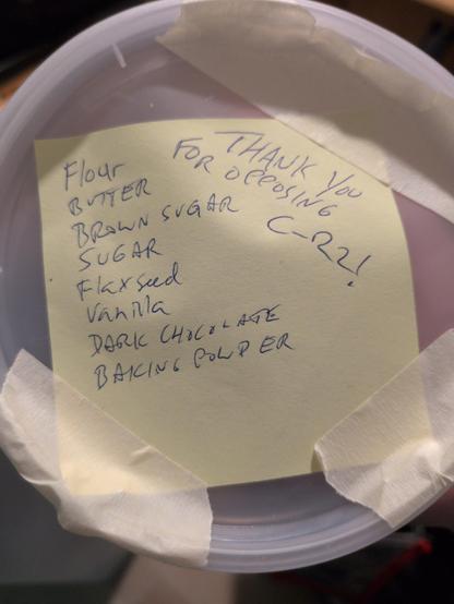 The top of a deli container full of cookies.  There is a post it note with the ingredients and "thanks for opposing c-22" written on it, held down on three corners by masking tape.