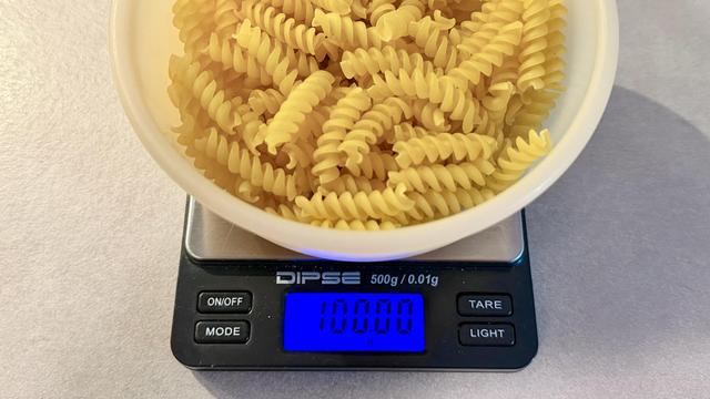 A bowl of raw pasta on a precision scale. The scale’s digital display shows 100.00 g, accurate to two decimal places.