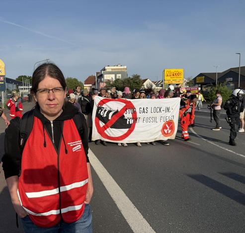 Ich gehe vor einer Demo, die eine Straße entlang zieht. Auf dem Transpi steht Stop Gas Lock In Fight fossil fuels. Ich trage eine rote Weste auf der paralemtarische Beobachtung steht.
