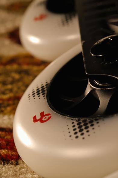 a tight shot of a white skate wheel with black halftone patterns and red letters that say 'UC' mounted on a black skate chassis