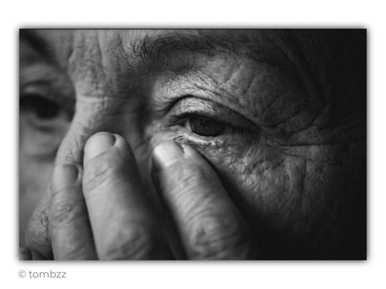 Analog black-and-white photograph showing an extreme close-up of part of an elderly woman’s face. The frame focuses mainly on her eyes and the hand pressed against her face. Her fingers partially cover her nose and cheek, naturally leading the viewer’s attention toward the right eye placed at the center of the image. The skin is deeply lined, marked with wrinkles and fine folds. Details of the skin texture, thin eyebrows, and delicate eyelashes remain clearly visible.
Her gaze appears tired and dimmed, yet still attentive. The second eye fades softly out of focus due to the very shallow depth of field. The lighting is soft and side-lit, emphasizing the texture of the face and the contrast between illuminated skin and the dark background. The photograph feels raw and intimate, built almost entirely on detail, texture, and the quiet emotion carried in the woman’s expression.